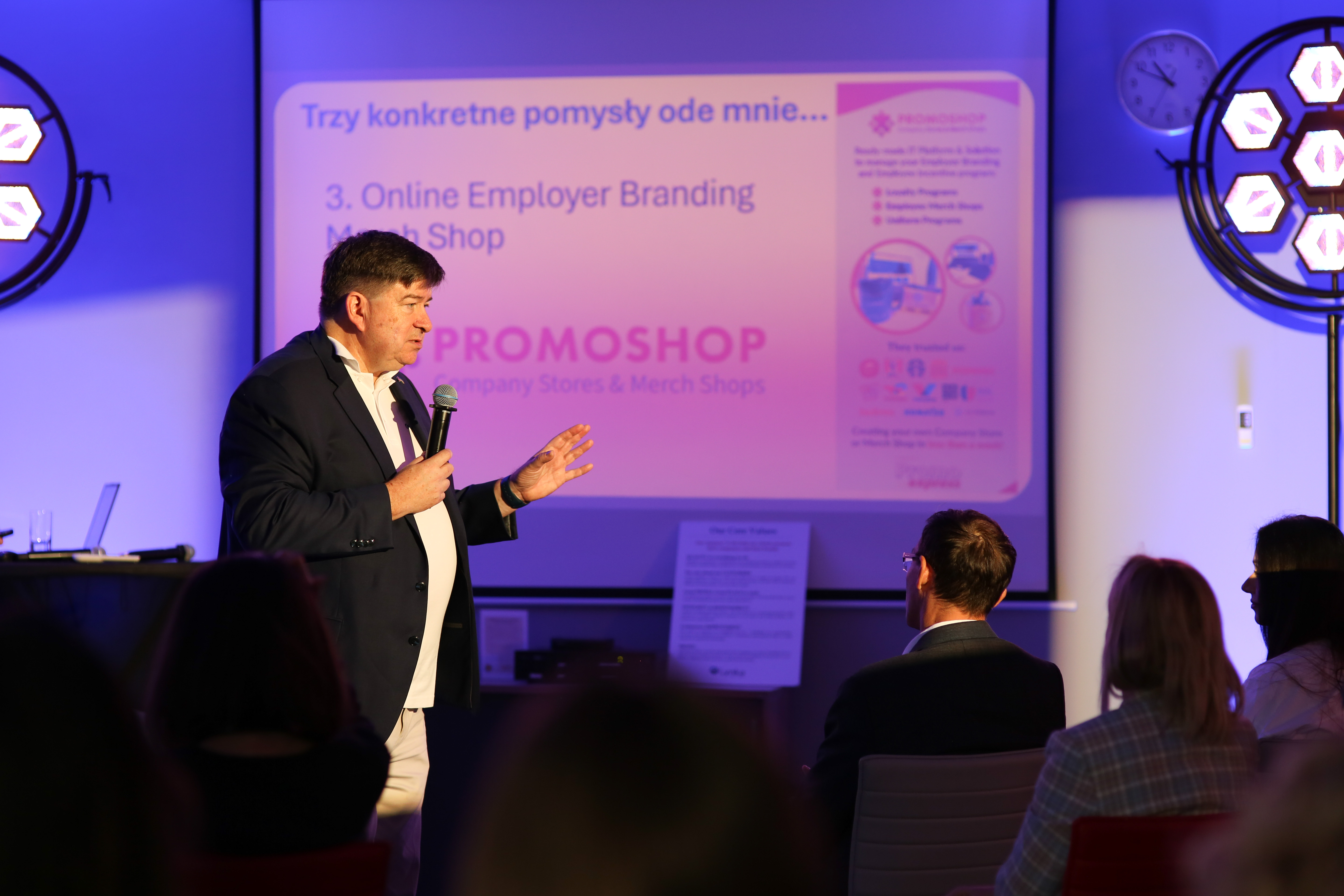 Katarzyna Luther at an Employer Branding conference, presentation on stage with microphone, clothing display of T-shirts and jackets in the background.