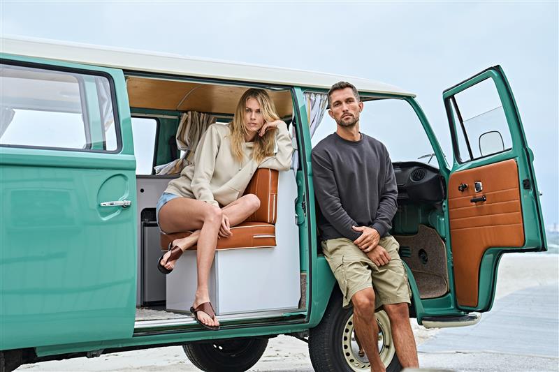 Man and woman wearing Tee Jays Label Free apparel posing by a green van on the beach.