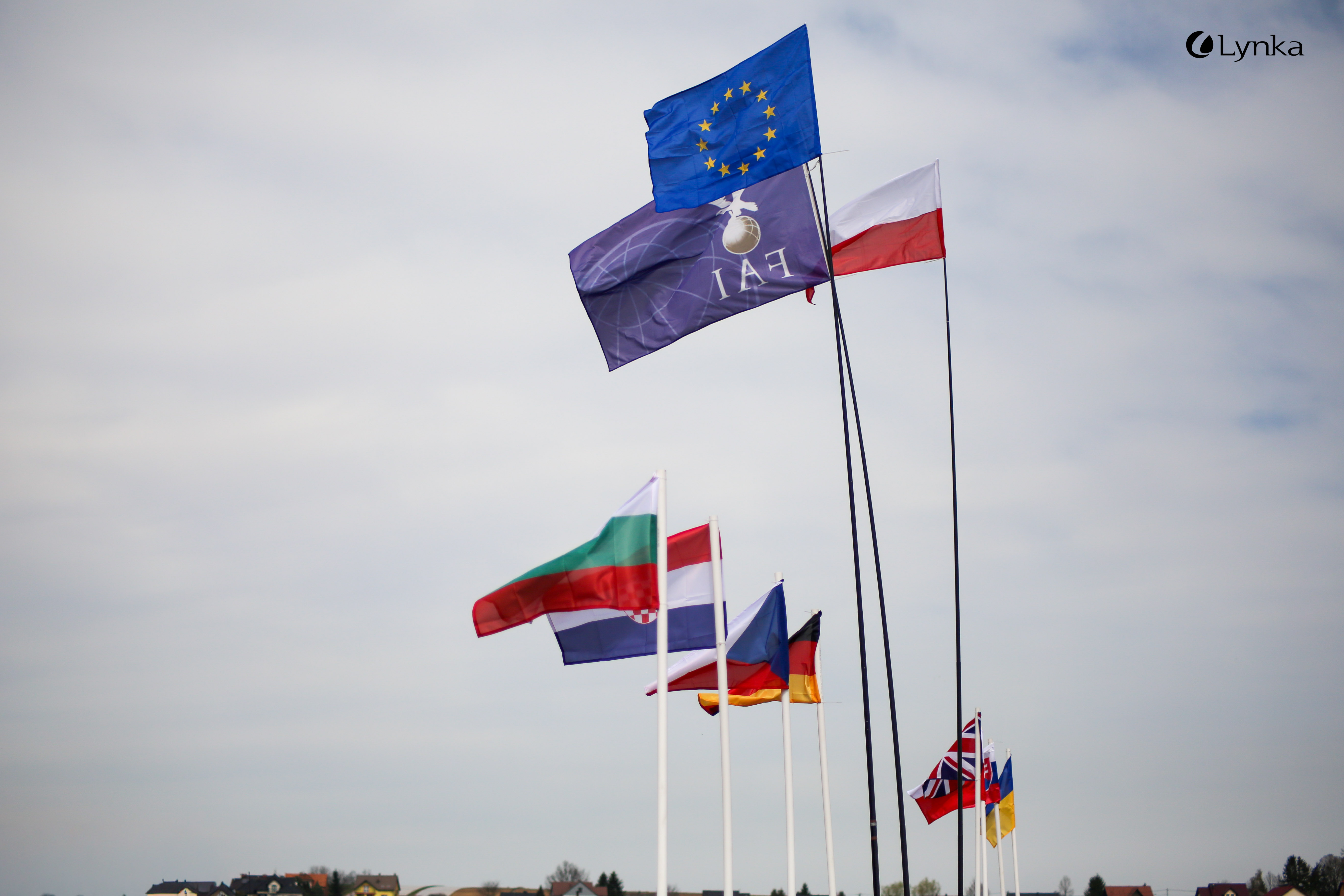 International flags, including EU, FAI, and Poland, flying on poles during a modeling competition against a cloudy sky.