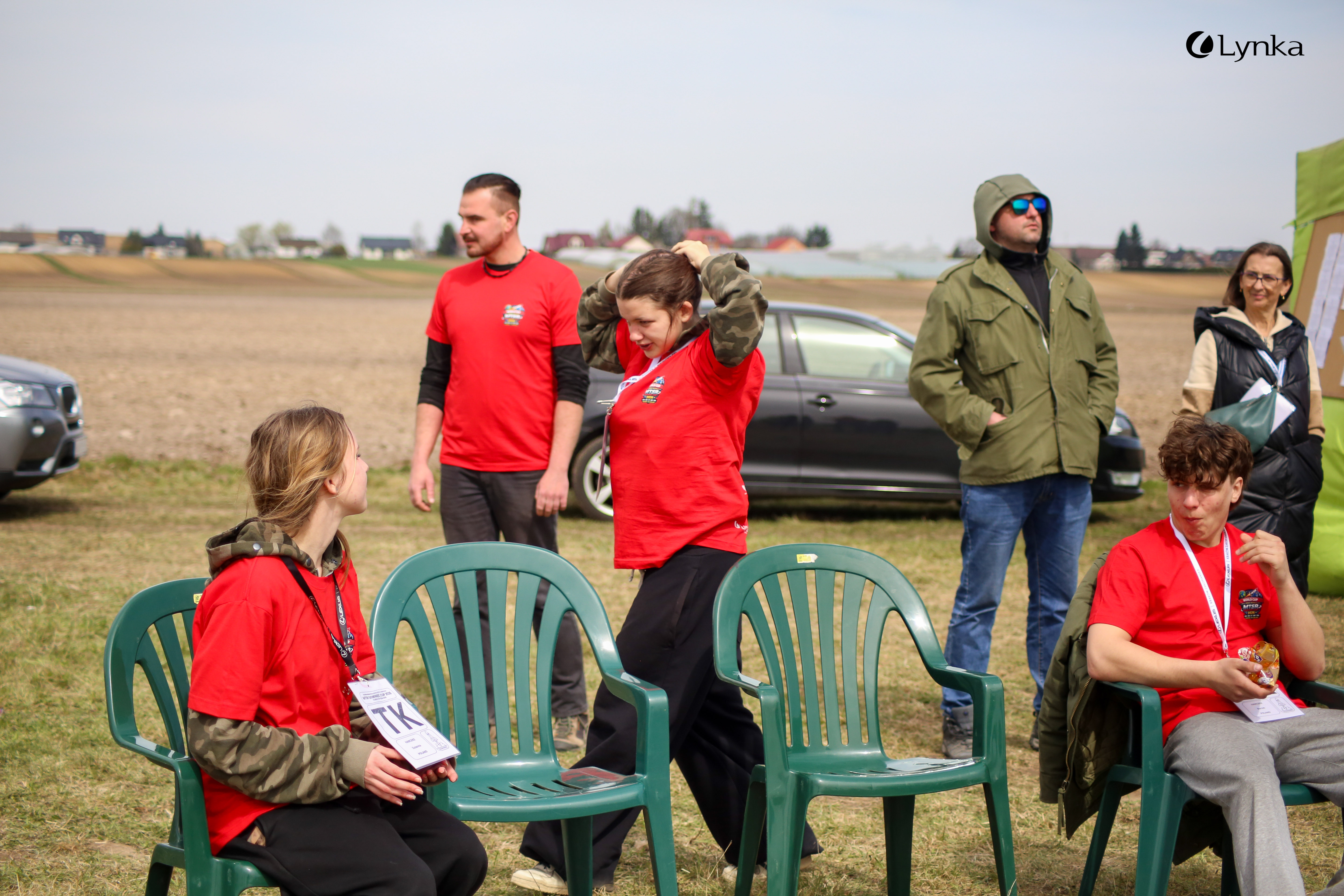 A group of young people and adults on a grassy field during a competition. Several people are sitting on green plastic chairs.