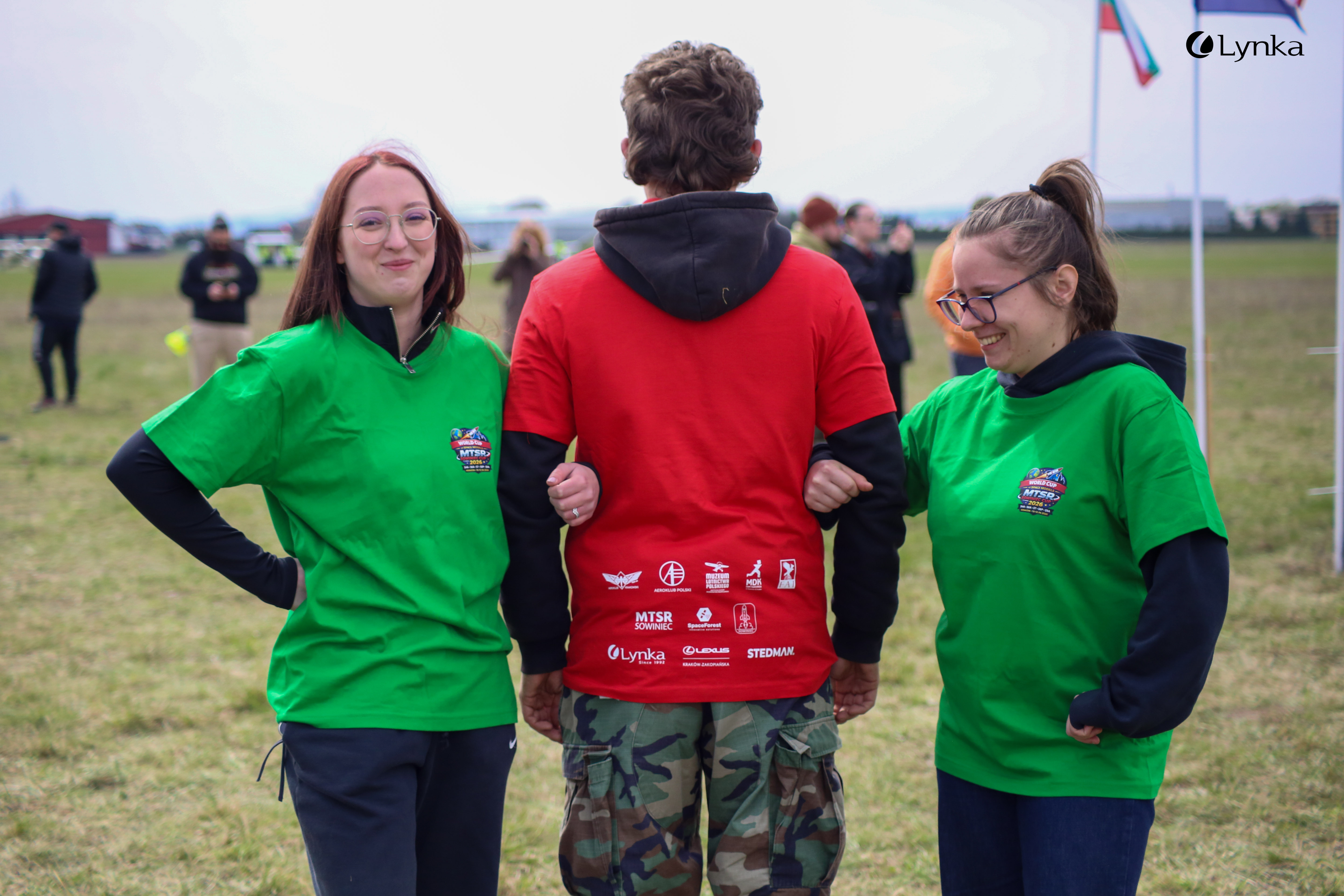 Three people presenting event t-shirts with MTSR 2024, Lynka, and Stedman logos. Two women in green and a man in a red shirt.
