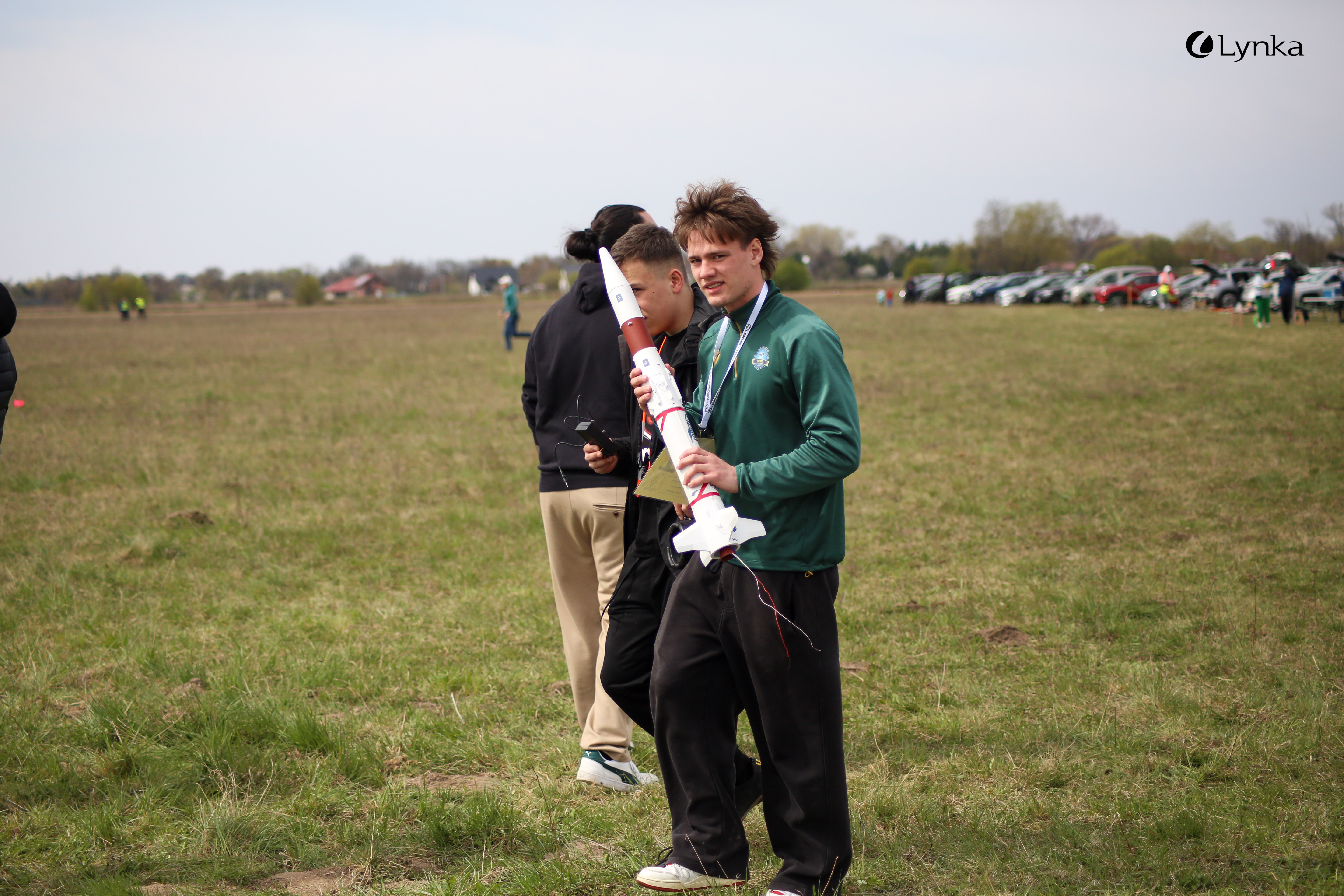 A young man in a green sweatshirt walks across a field, carrying a red and white model rocket. A row of parked cars is visible in the background.