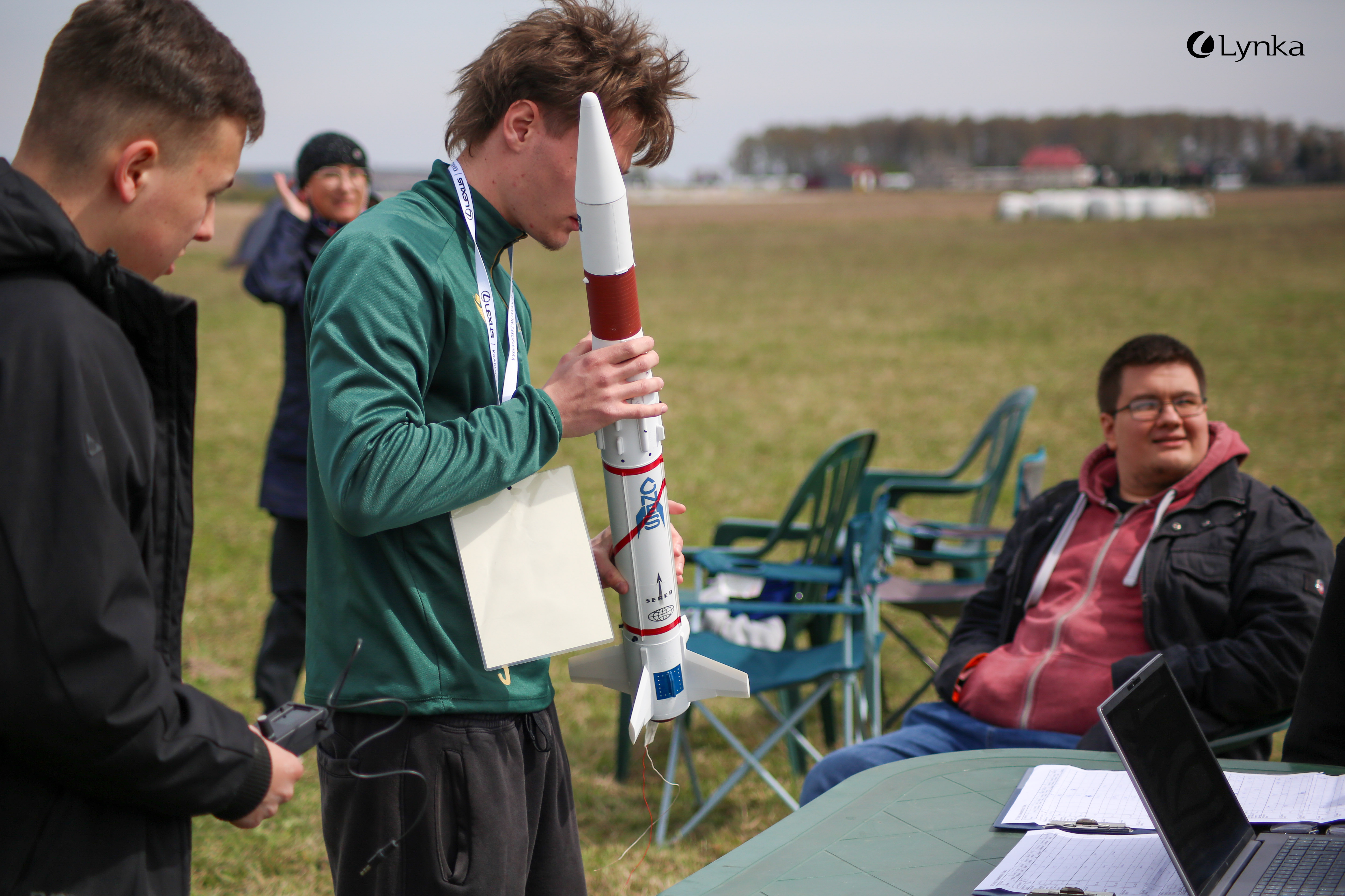Participant holding a white and red model rocket for inspection at the competition, next to a table with a laptop.