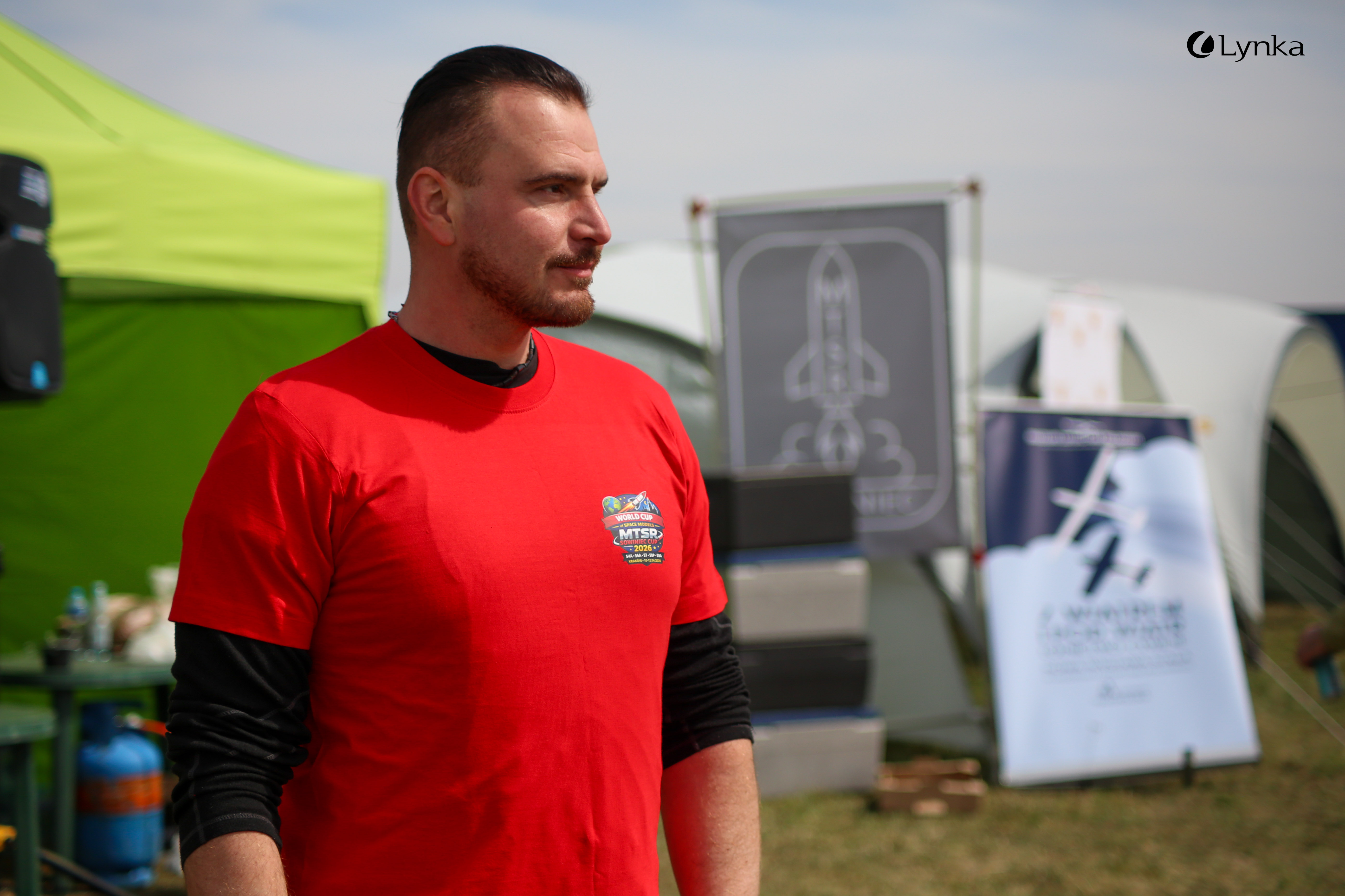Man in a red event T-shirt with the "World Cup of Space Models MTSR 2026" logo on the chest at an outdoor space modeling competition.