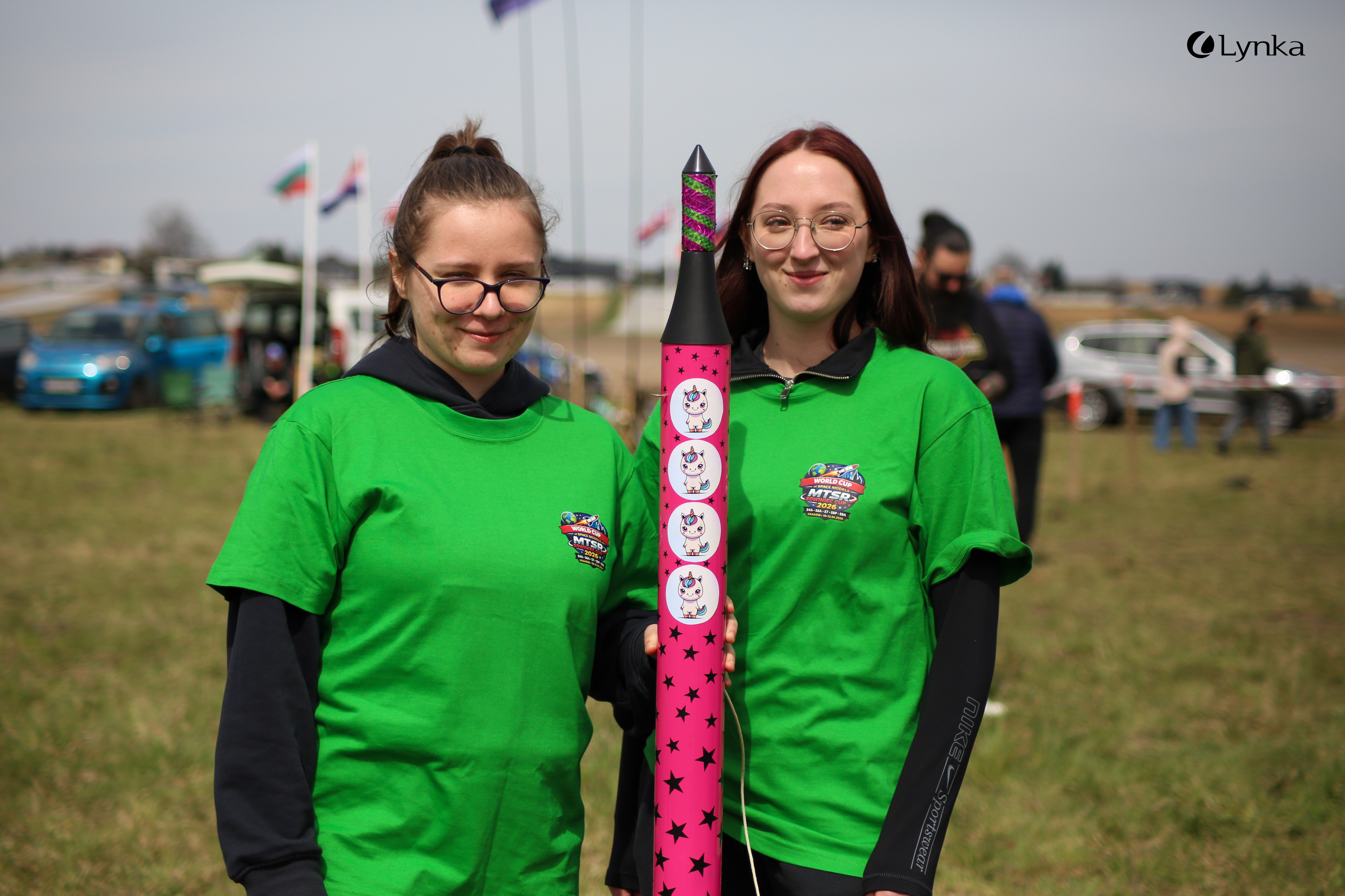 Two women in green event T-shirts smiling and holding a pink rocket decorated with unicorn stickers.