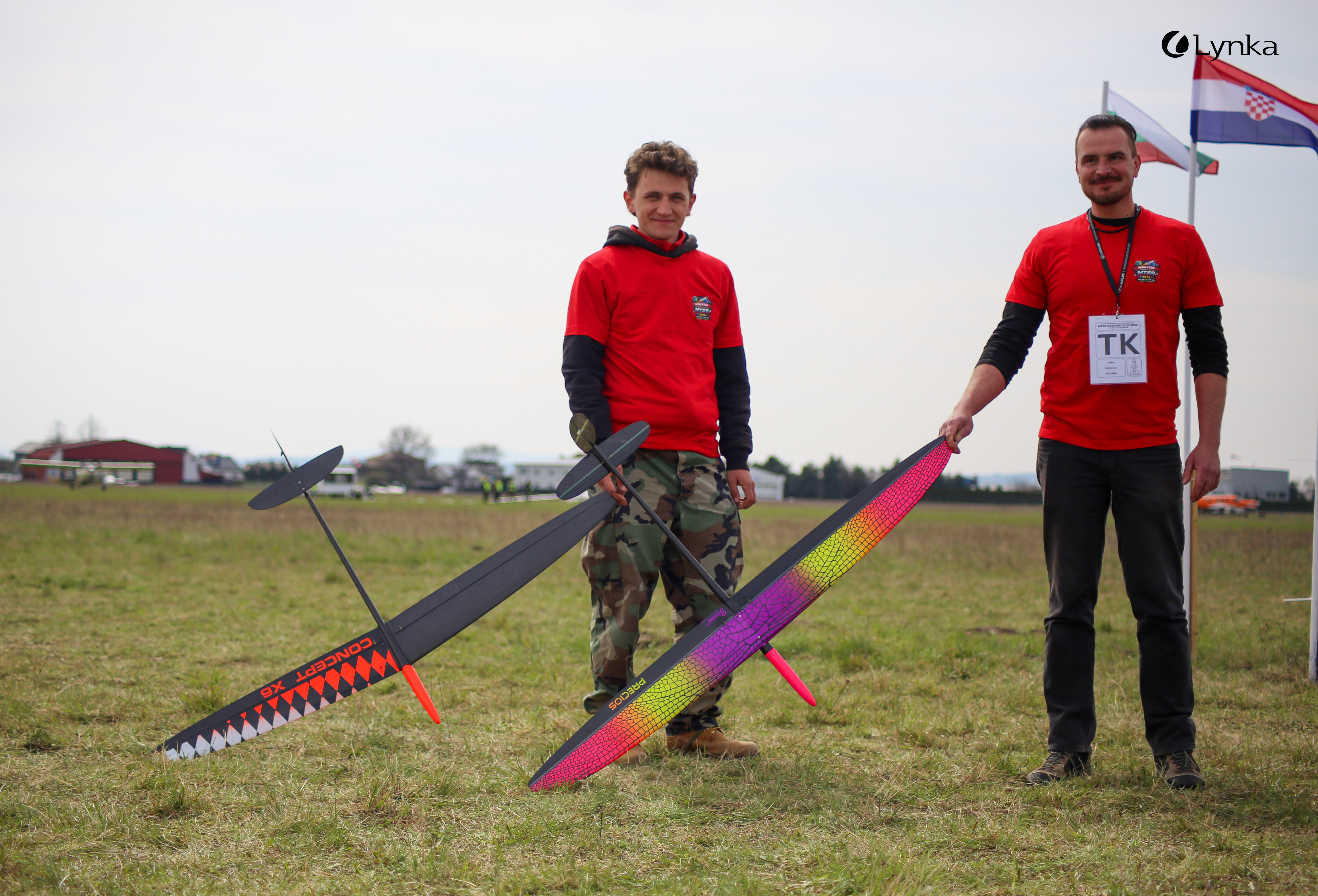 Two men in red t-shirts standing on a grass field, holding large, modern model gliders with colorful wings.