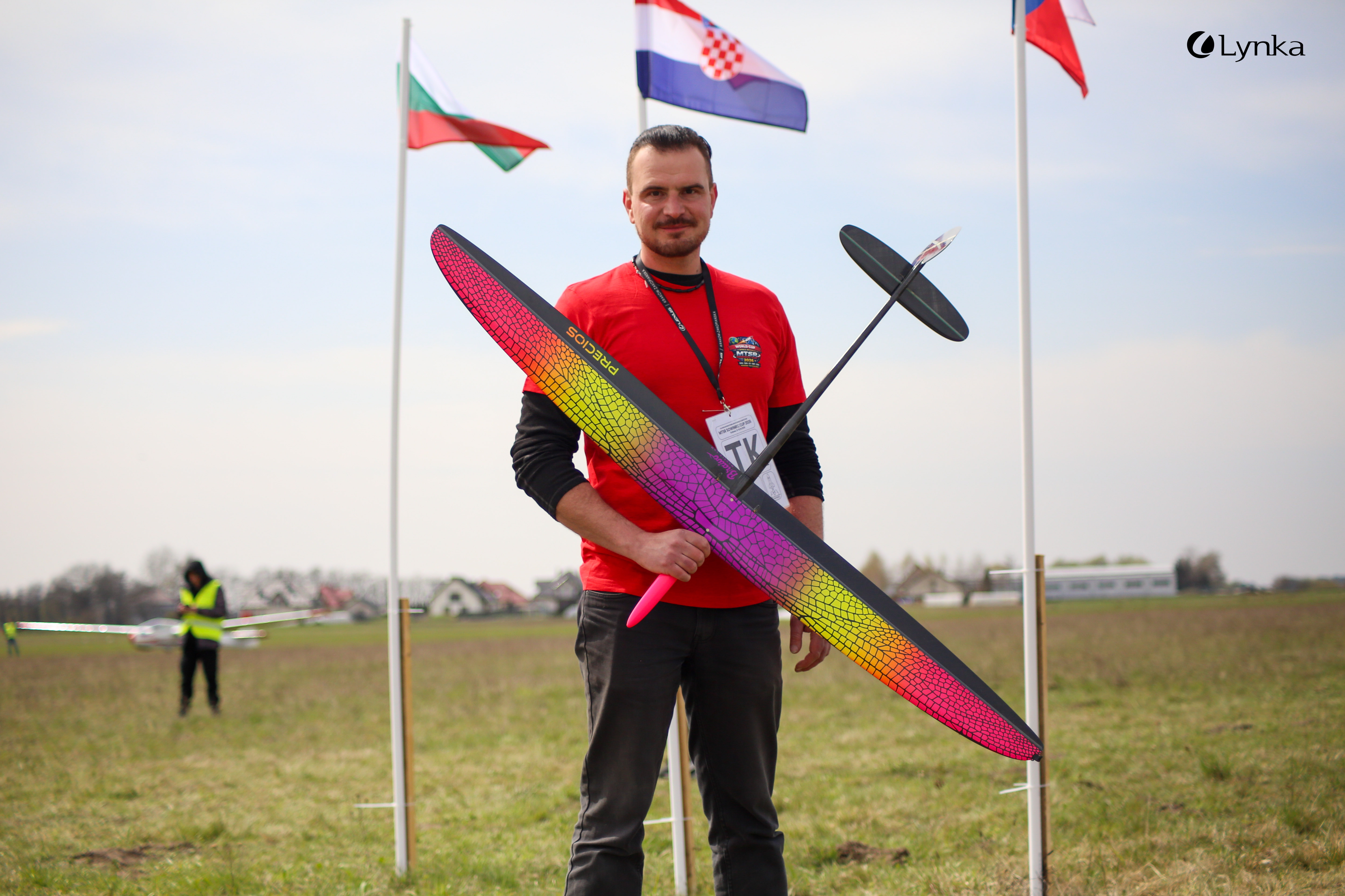 A man in a red t-shirt poses for a photo on a grassy field, holding a large model glider with a bright, colorful wing.