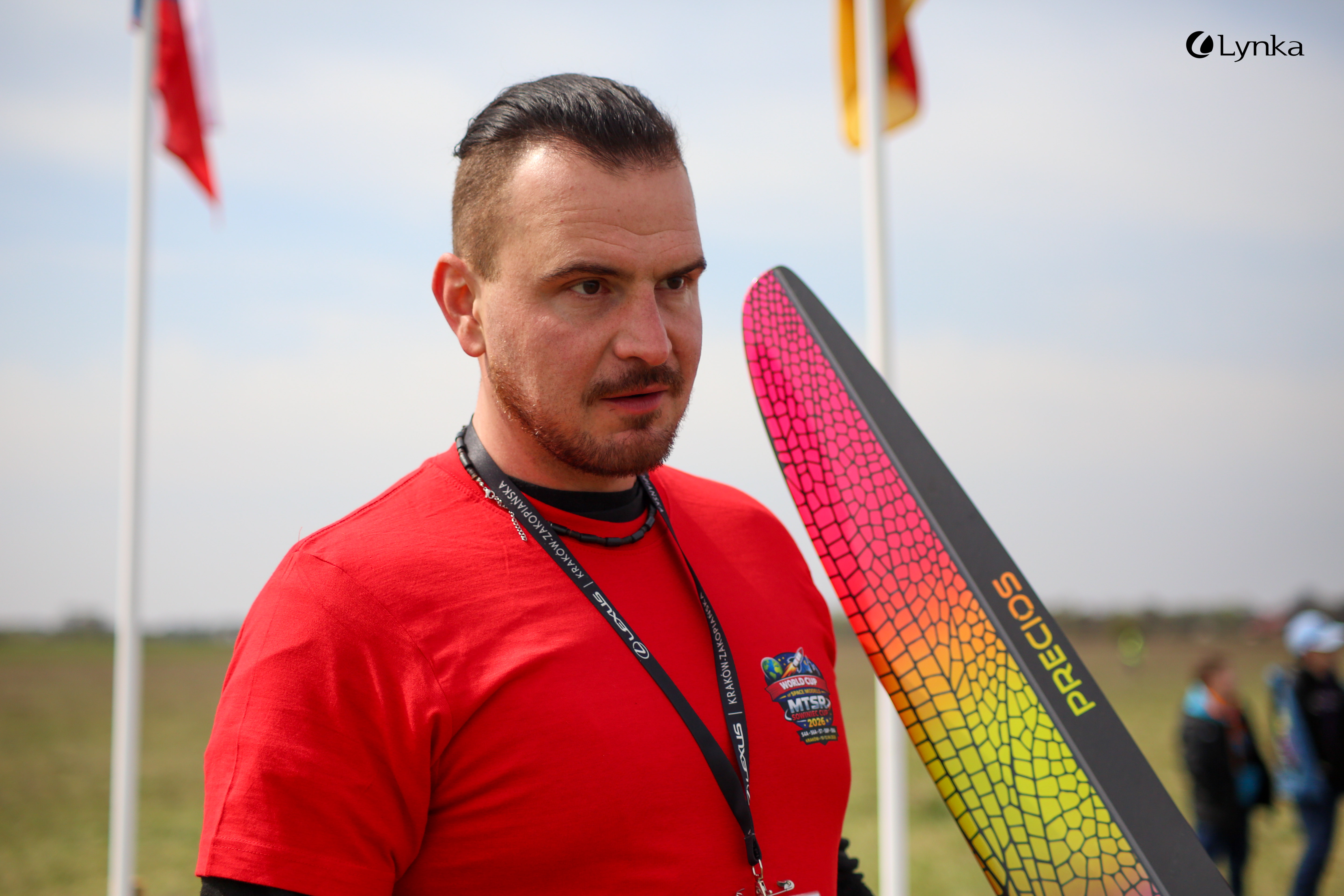 A portrait of a young man in a red MTSR 2024 t-shirt standing on a field. A section of a black model is visible next to him.