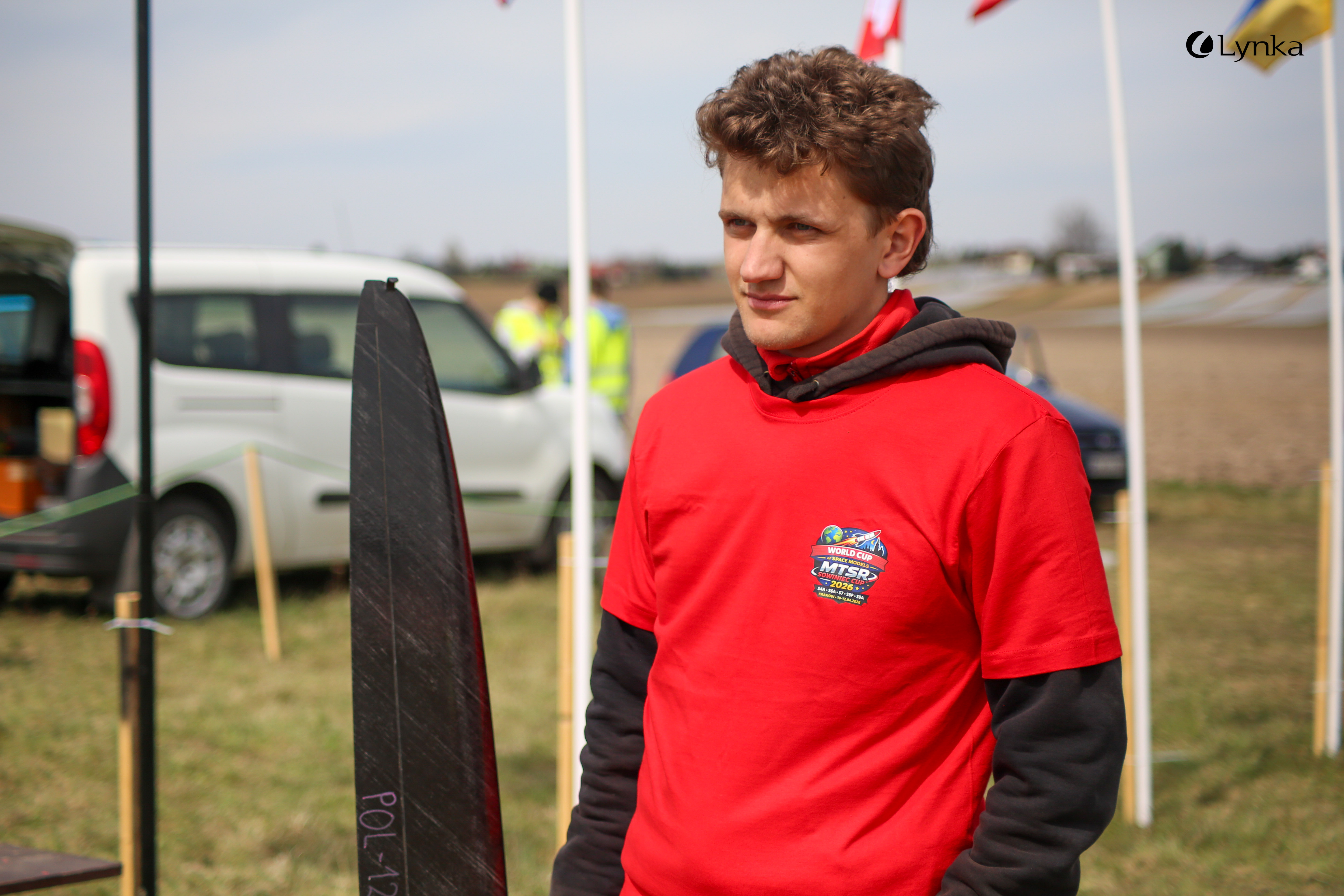 A portrait of a young man in a red MTSR 2024 t-shirt standing on a field. A section of a black model is visible next to him.