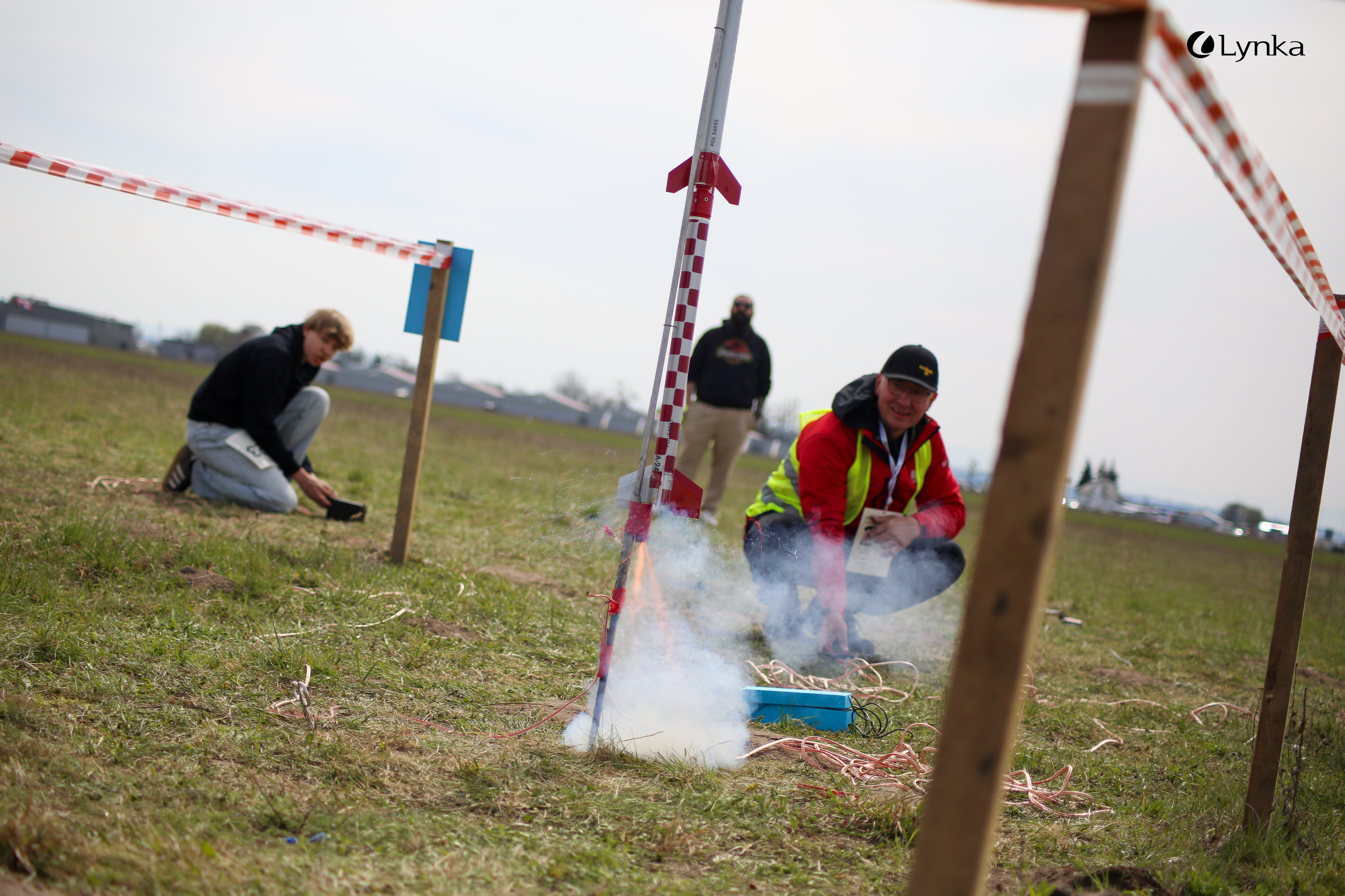 The moment of a model rocket launch with smoke and sparks at the base, shown from a low angle on the launch field.