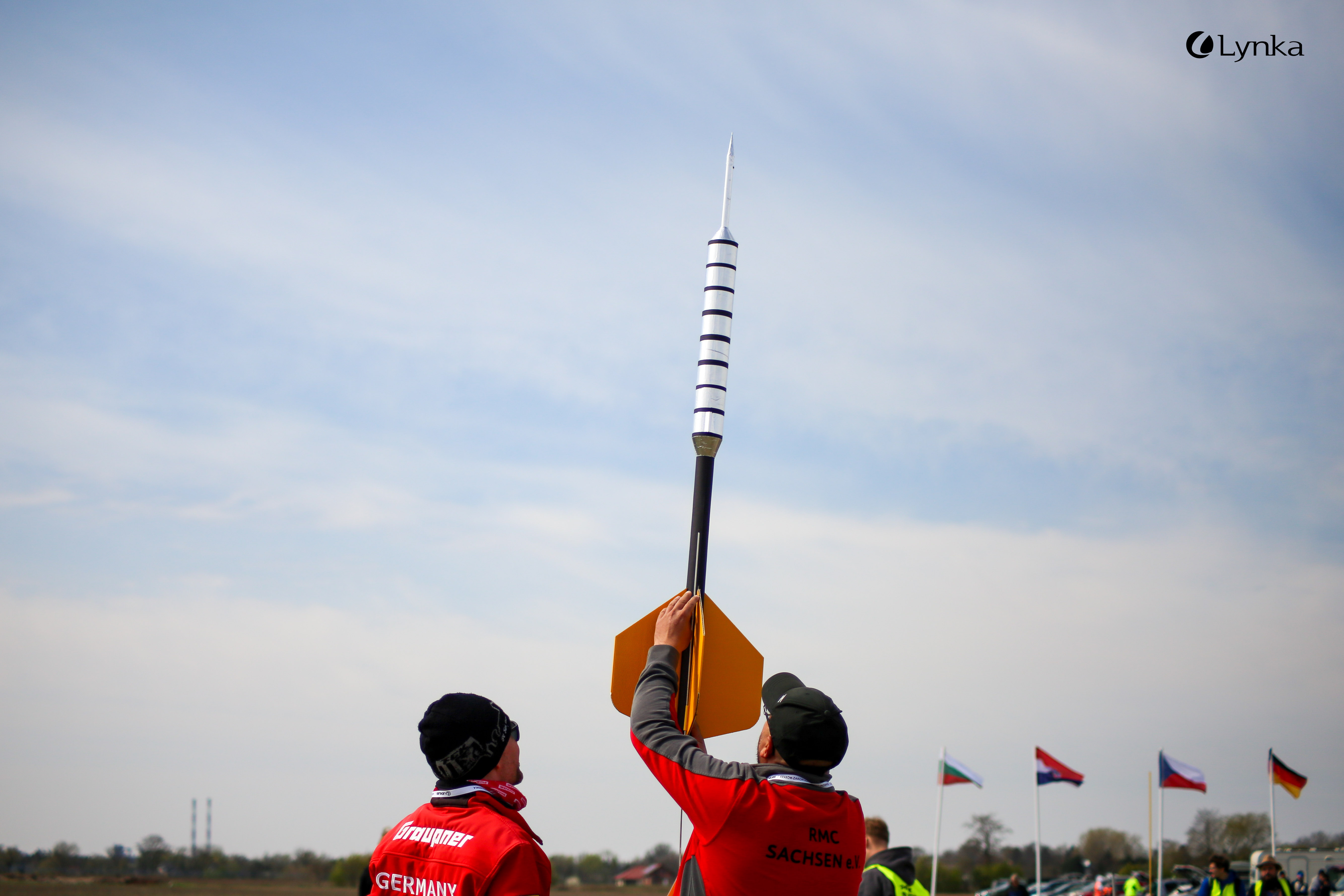 Two men in red jackets labeled "Germany" and "RMC Sachsen" holding a large silver and black model rocket vertically.