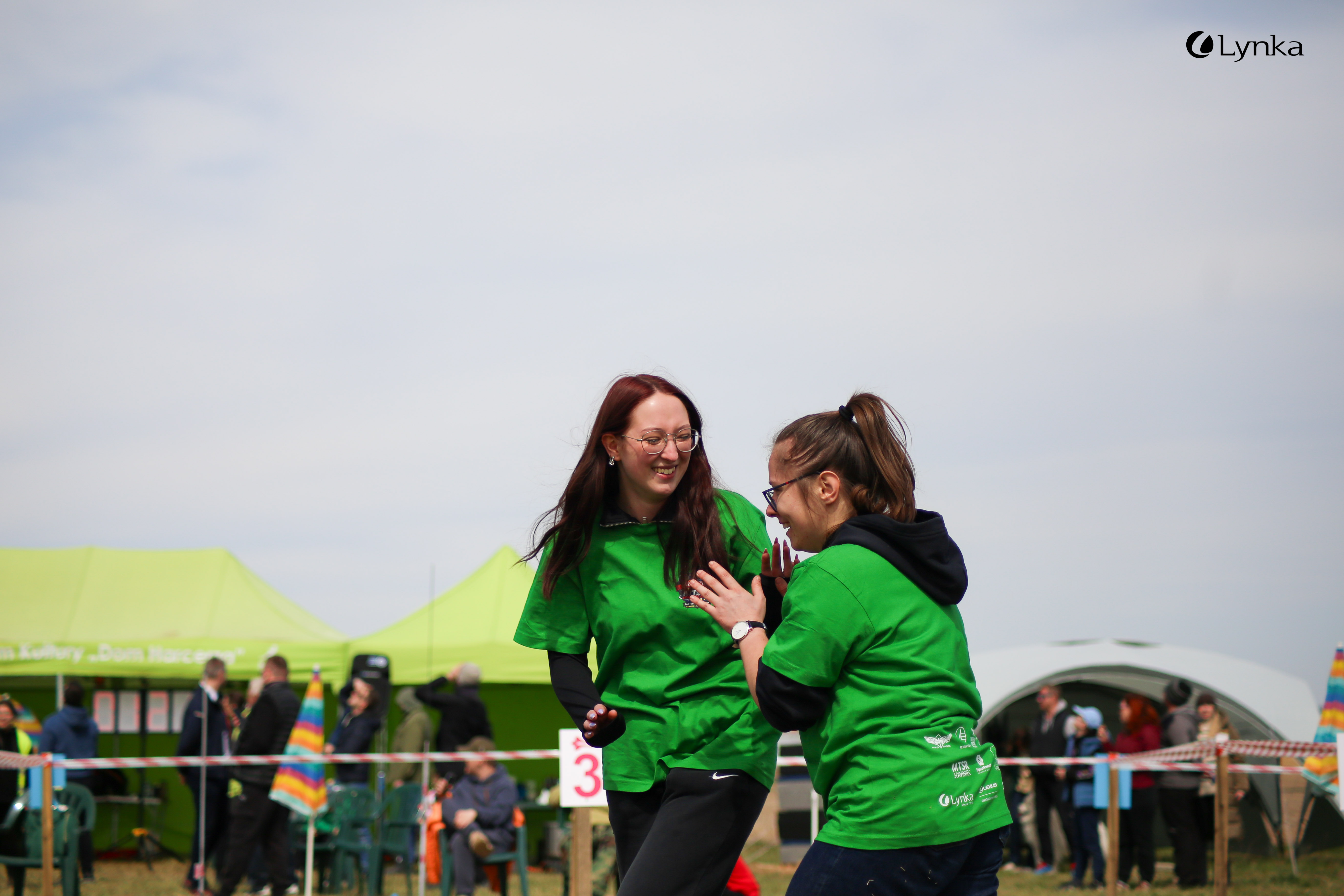 Two young women in green MTSR 2024 t-shirts laugh and talk on a grassy field on a sunny day