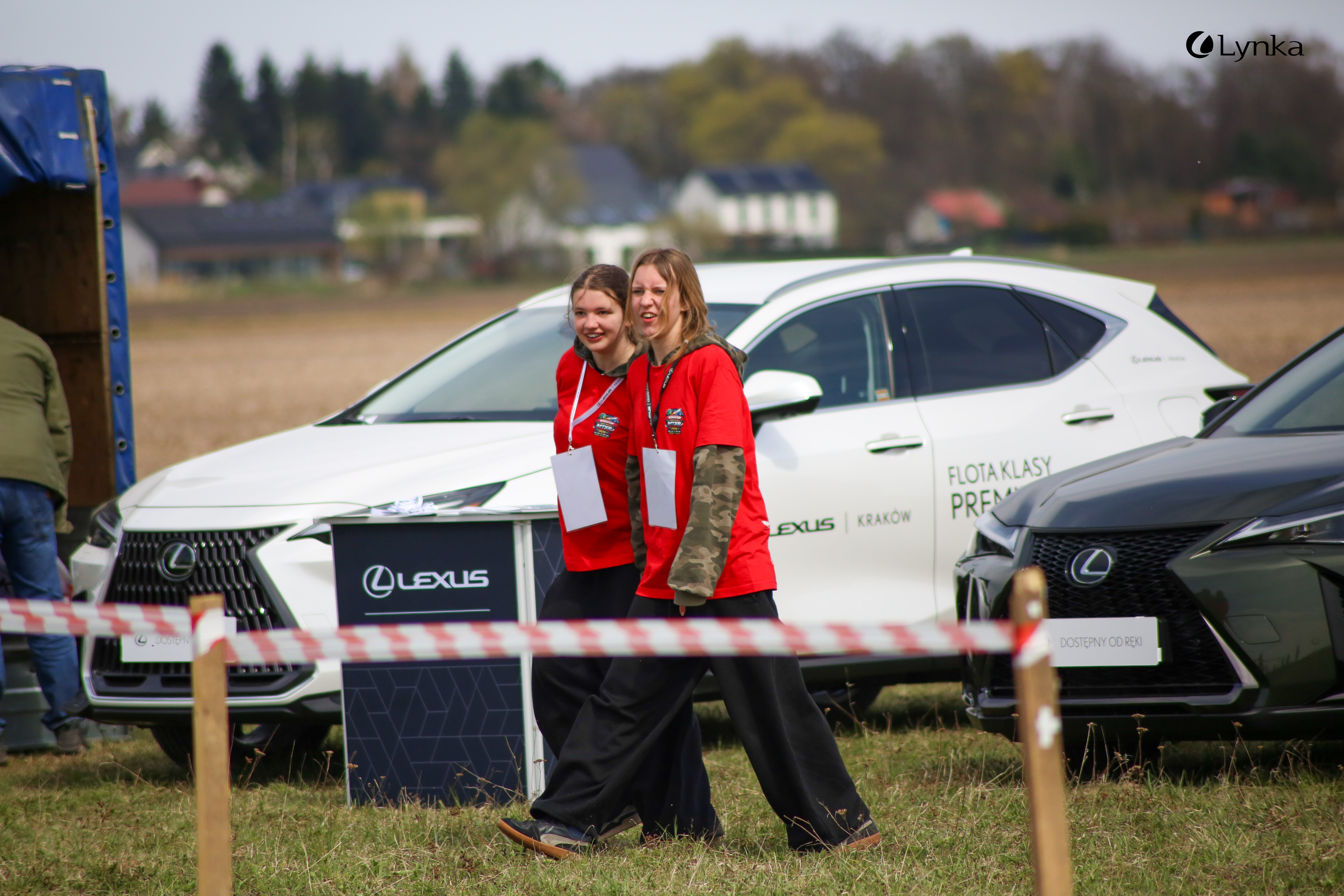 Two young women in red t-shirts walk across a grassy field. White and black Lexus cars and buildings are visible in the background.
