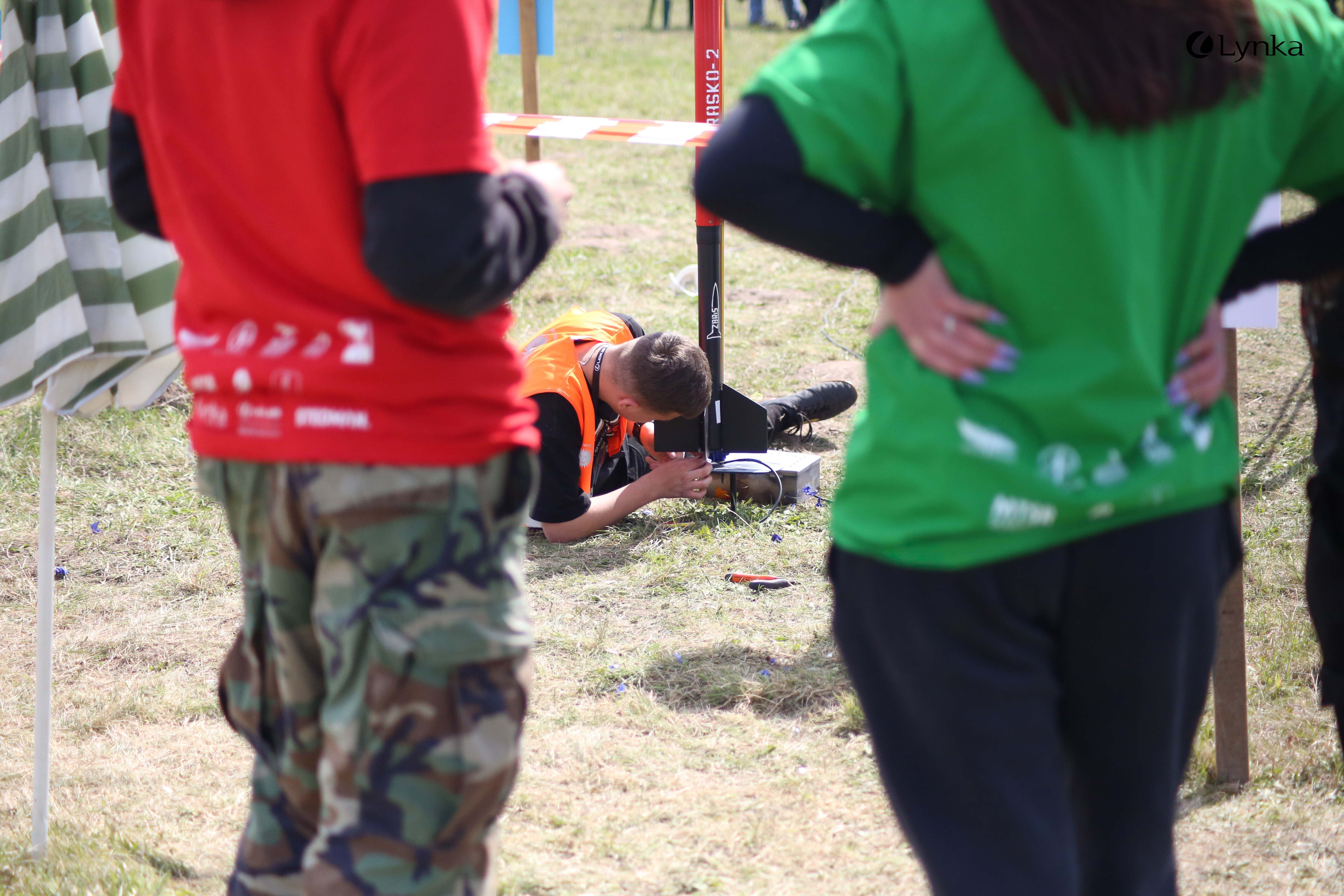 View from behind two people in t-shirts of a man in an orange vest kneeling on the grass and working on a model rocket.