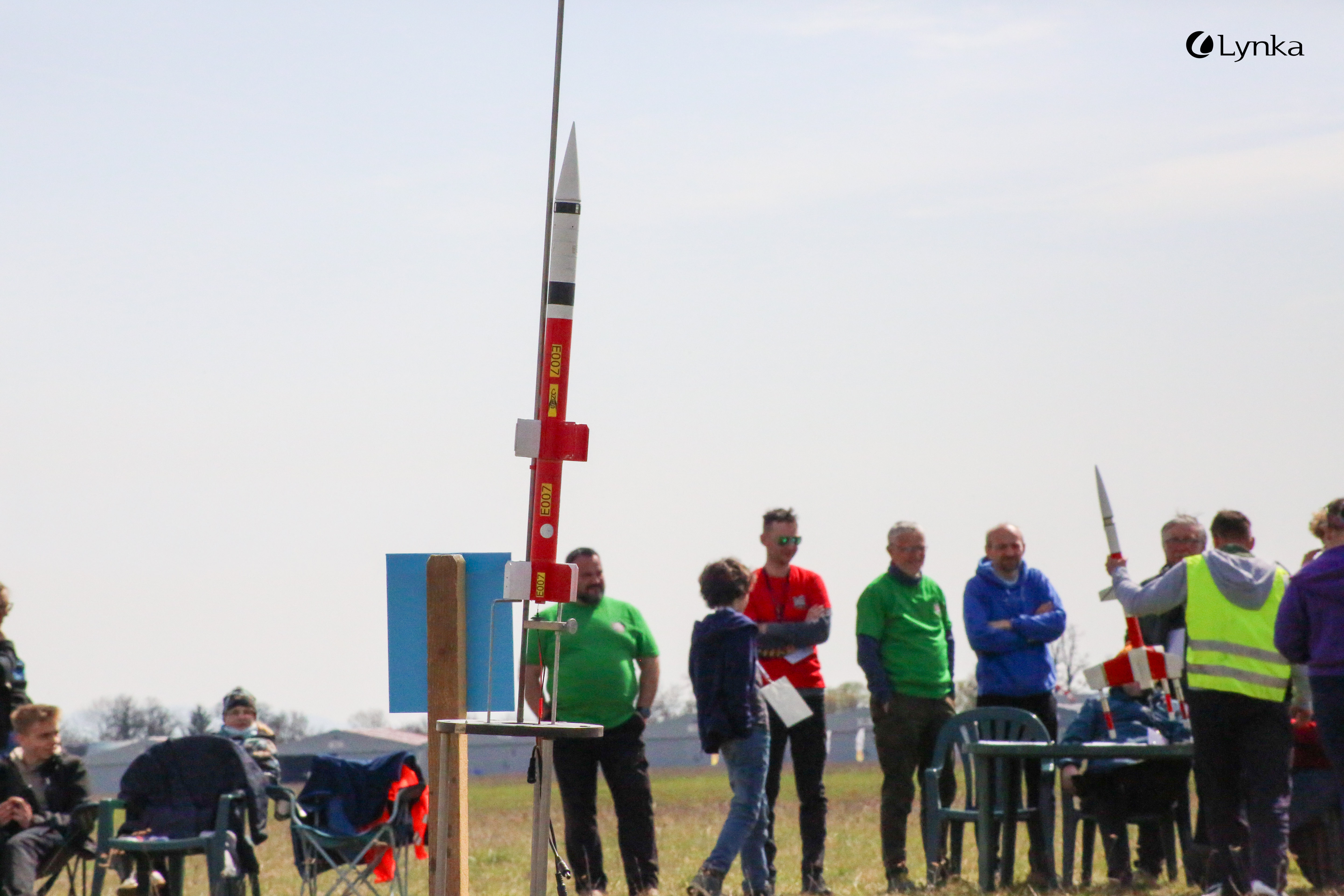 A red and white model rocket on a launch pad ready for takeoff. Competition participants are visible in the background on an open field.