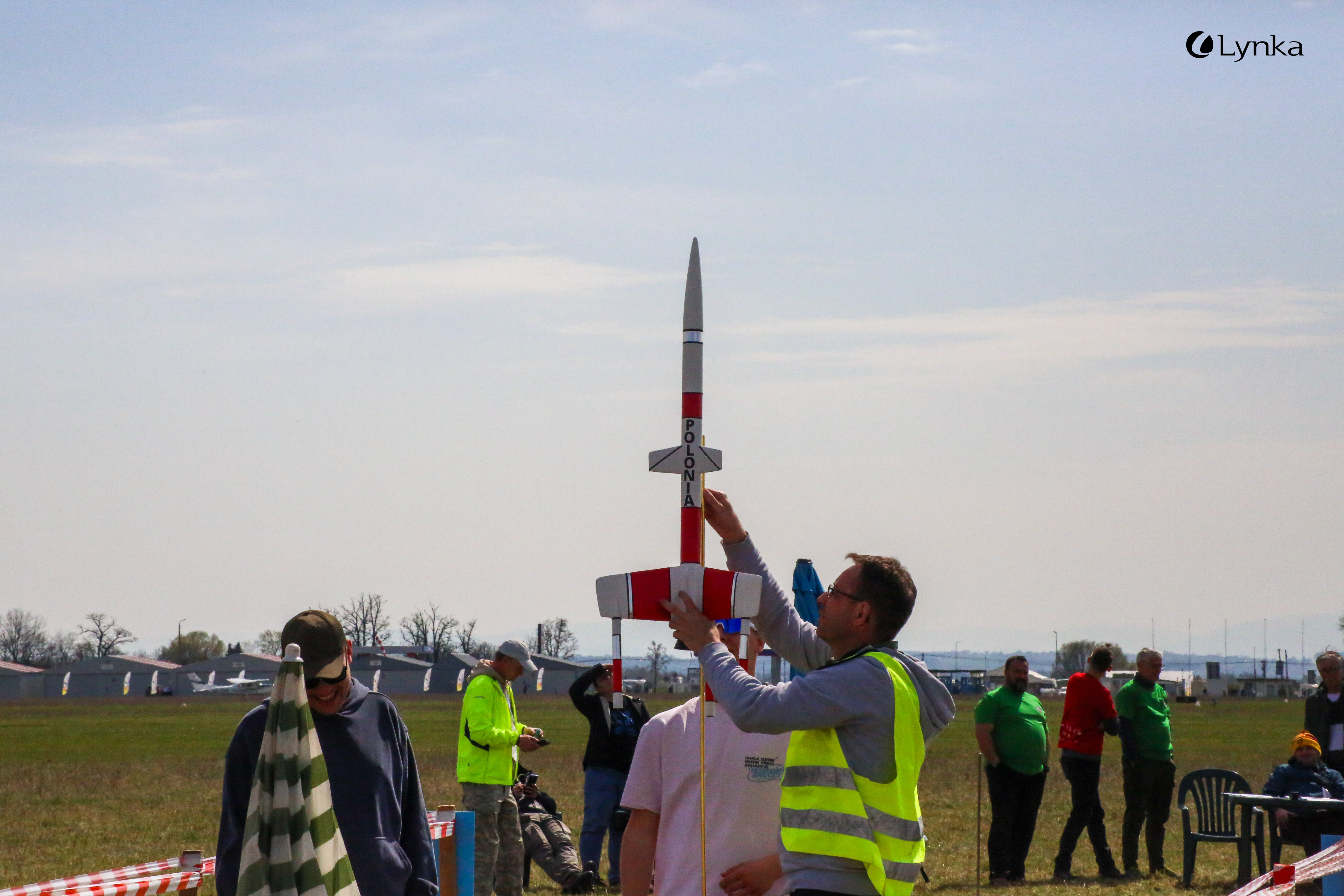 A man in a hi-vis vest adjusts a red and white model rocket marked "POLONIA" at the launch station during an outdoor event.