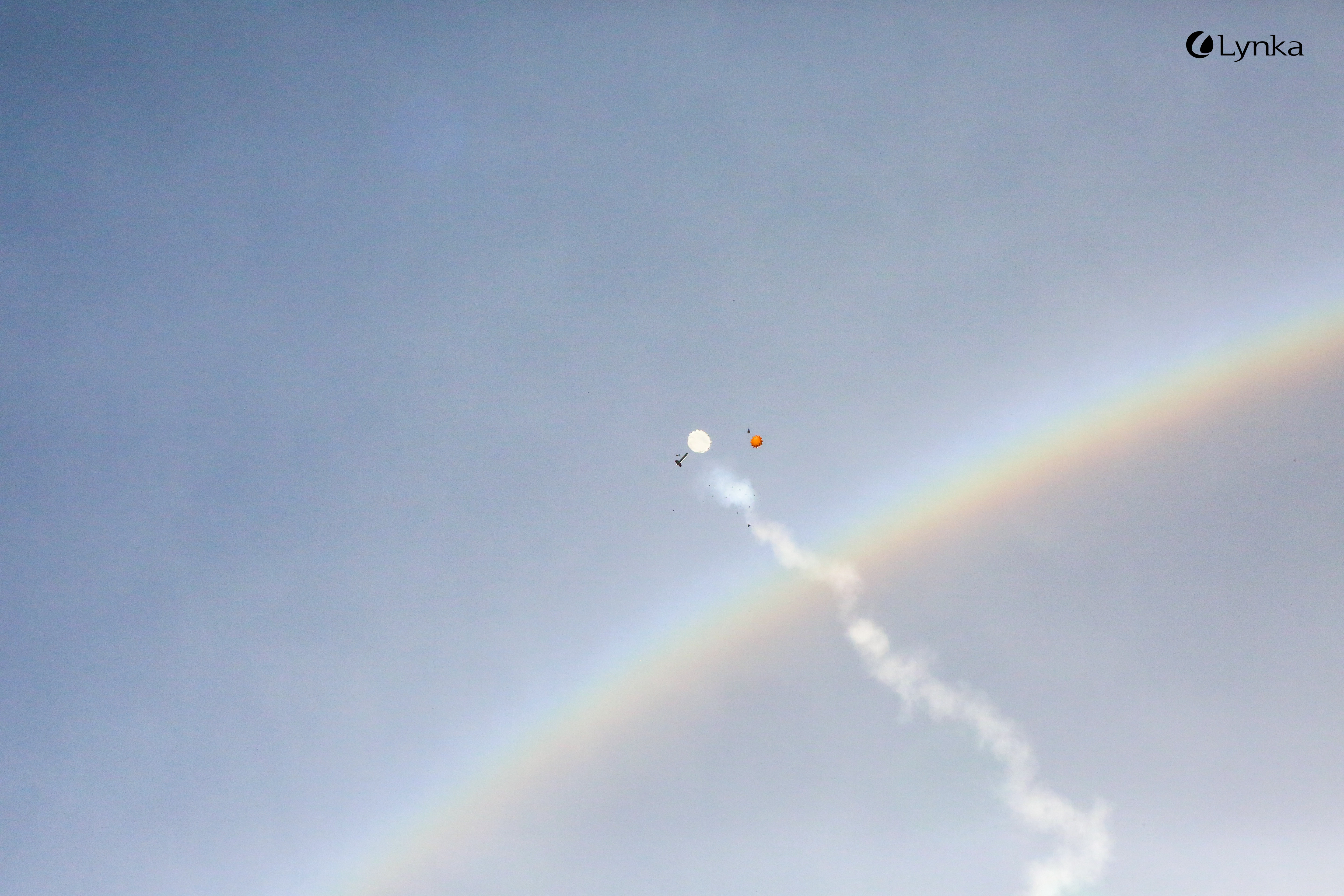 A model rocket descending on a white parachute, leaving a smoke trail in the sky next to a bright rainbow.