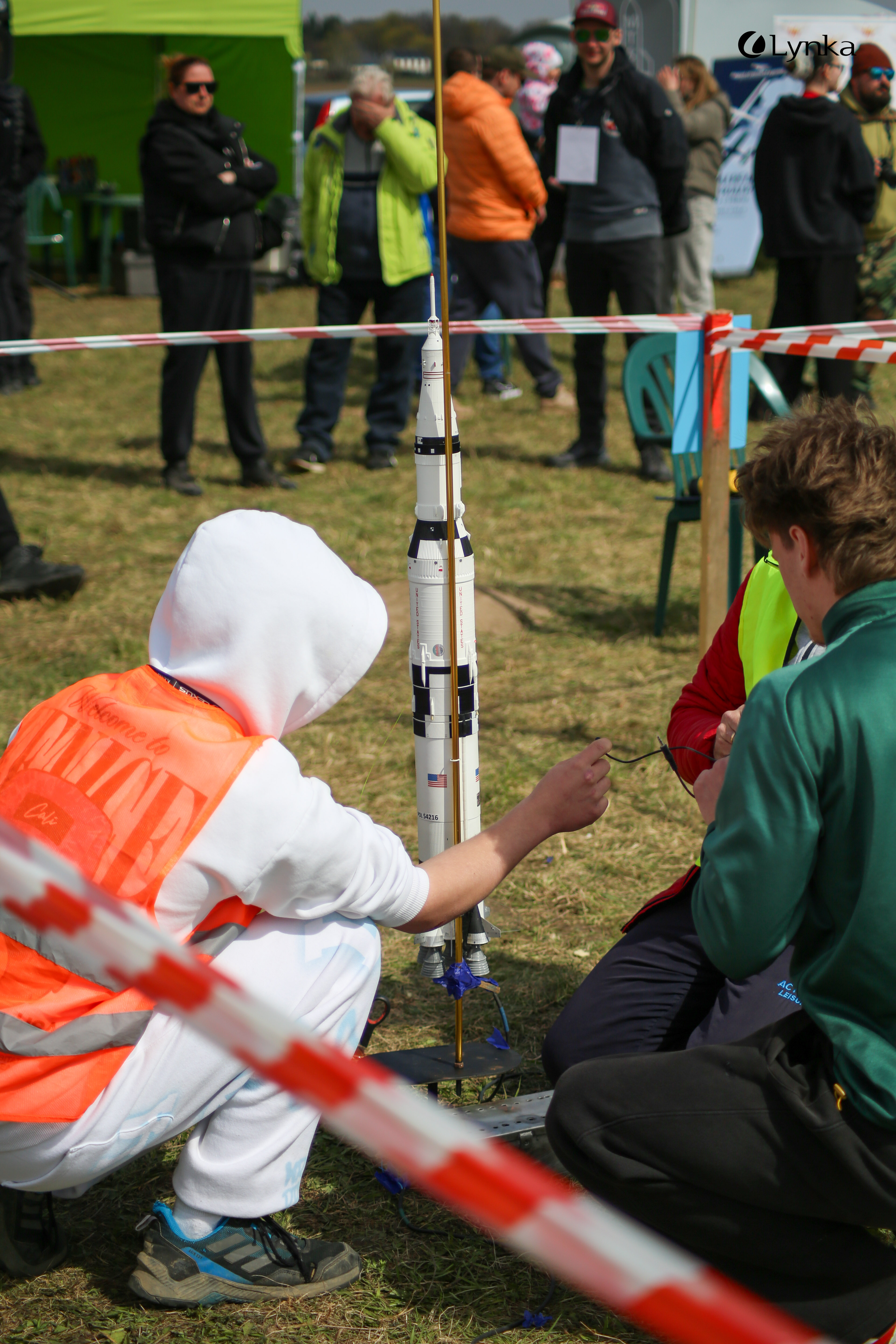 Launch preparations: a person in an orange vest kneels beside a detailed Saturn V rocket model on a grassy airfield.
