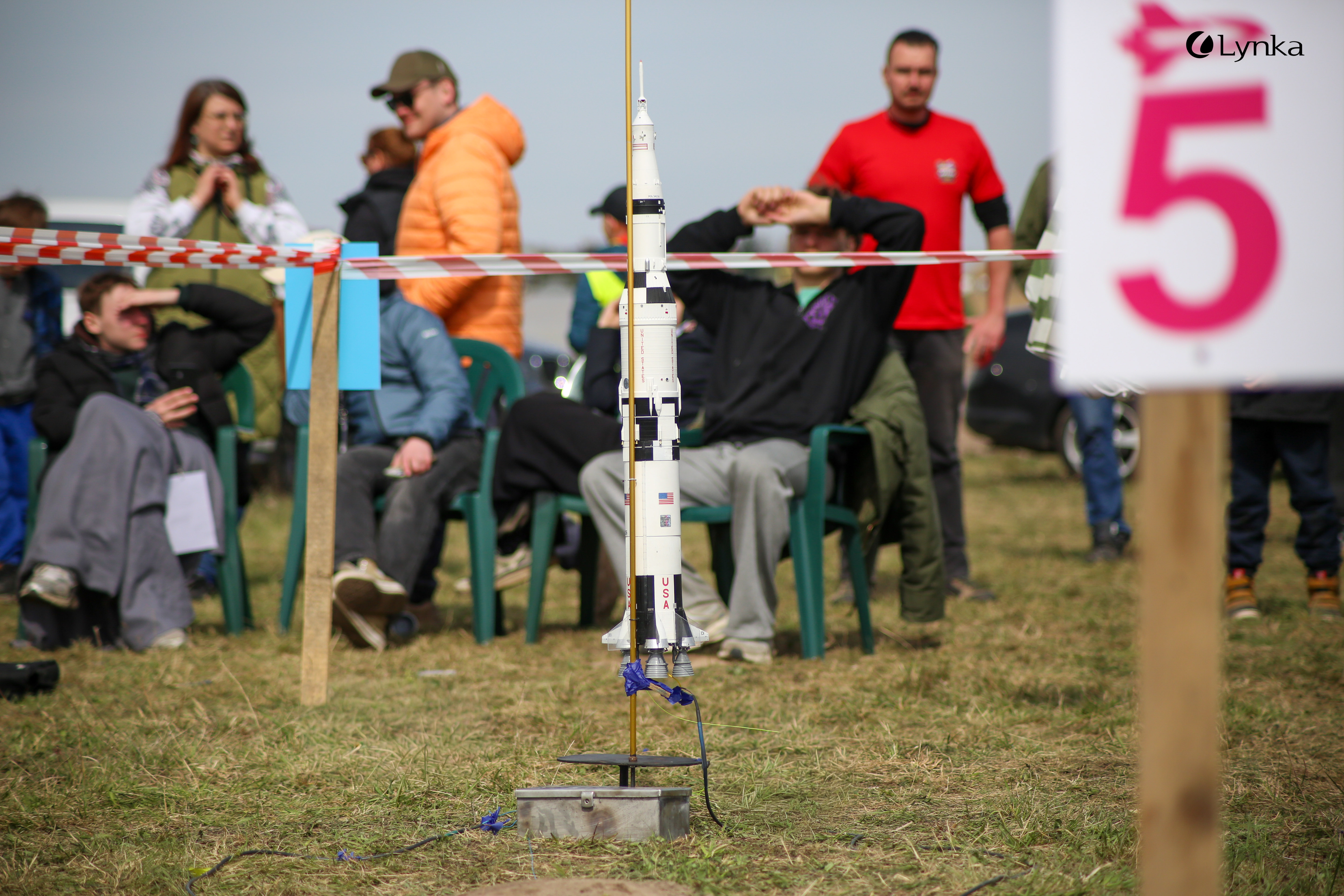 Detailed Saturn V model rocket on a launch pad during the MTSR 2026 competition. Participants sitting on the grass in the background.