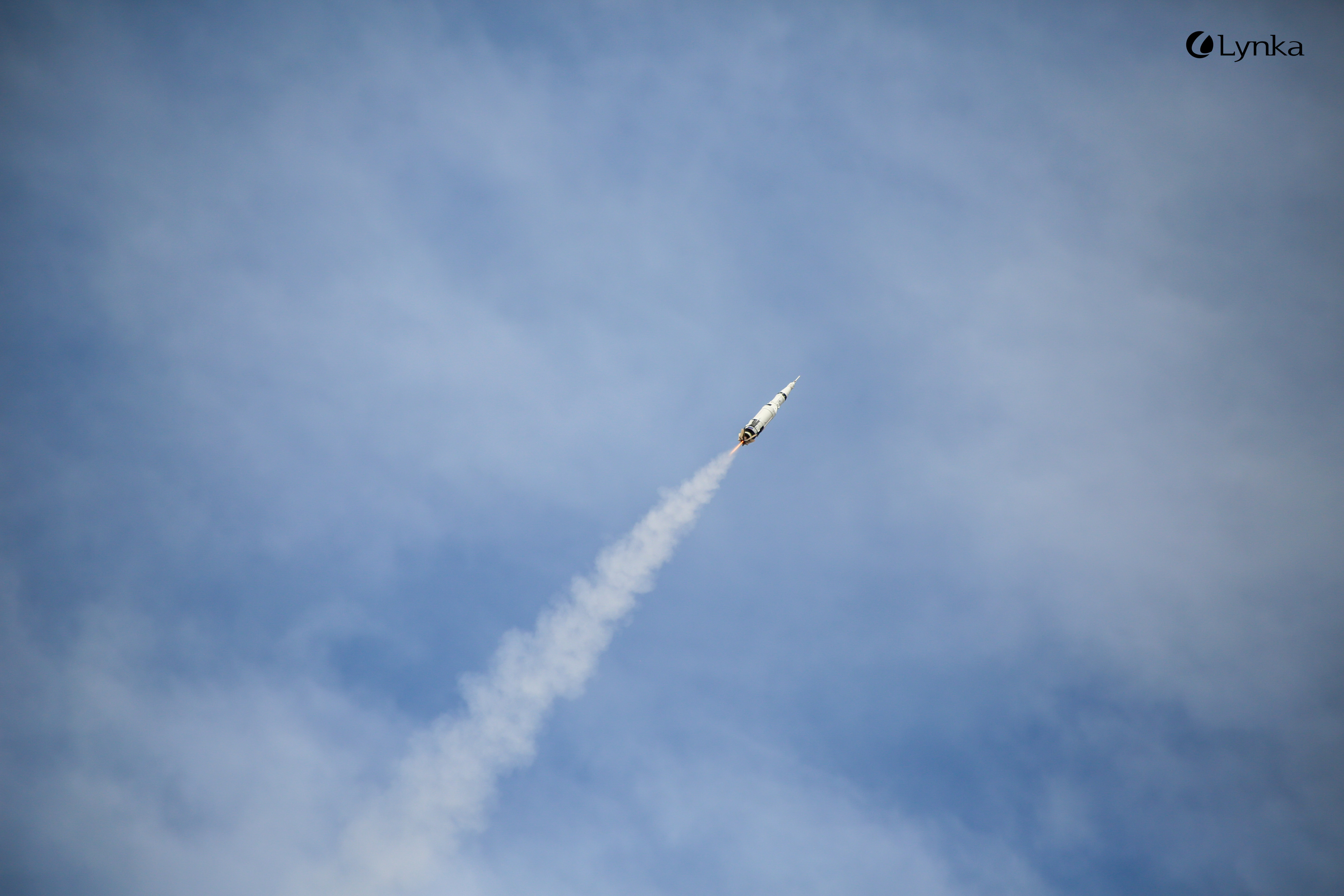 A model rocket ascends into a blue, partly cloudy sky, leaving a distinct smoke trail behind.