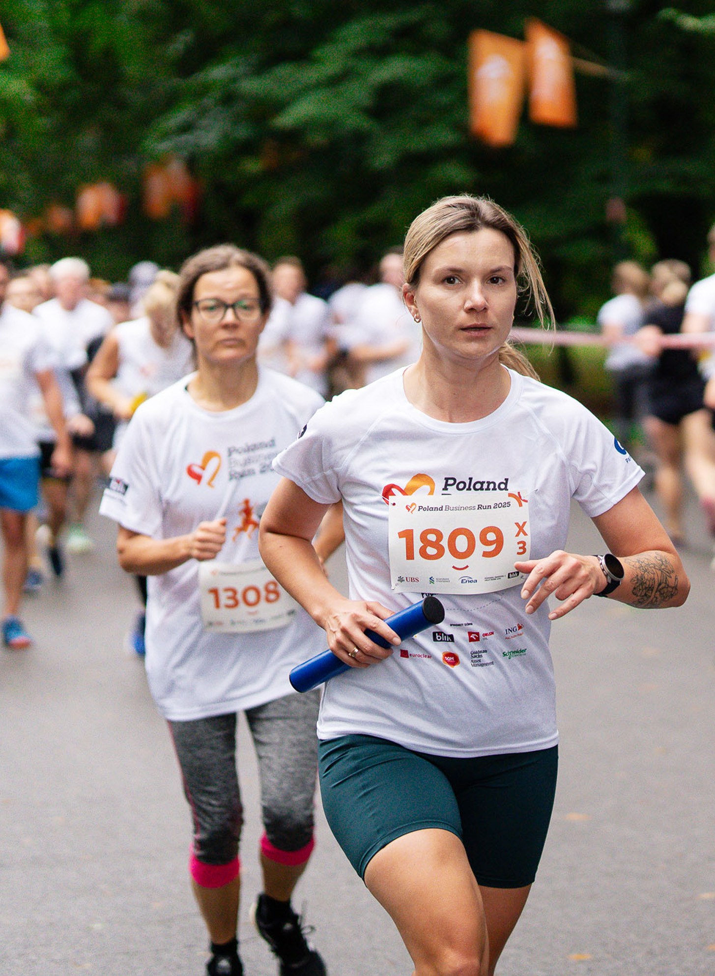 Biegaczka w białej koszulce Poland Business Run z numerem biegnie po parkowej alei, trzymając pałeczkę sztafetową.