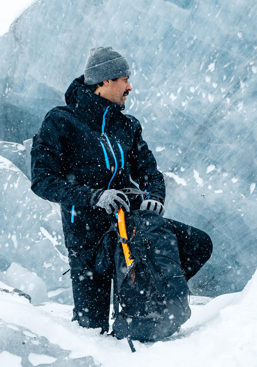 Man in a dark 3-in-1 jacket kneeling with an ice axe against a glacier wall.